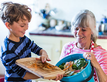 A woman holding a blue compost bucket while a young boy adds food scraps from a cutting board to it.